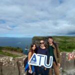 Three International Student Society members holding an ATU sign at the Cliffs of Moher, with the ocean and green cliffs in the background.