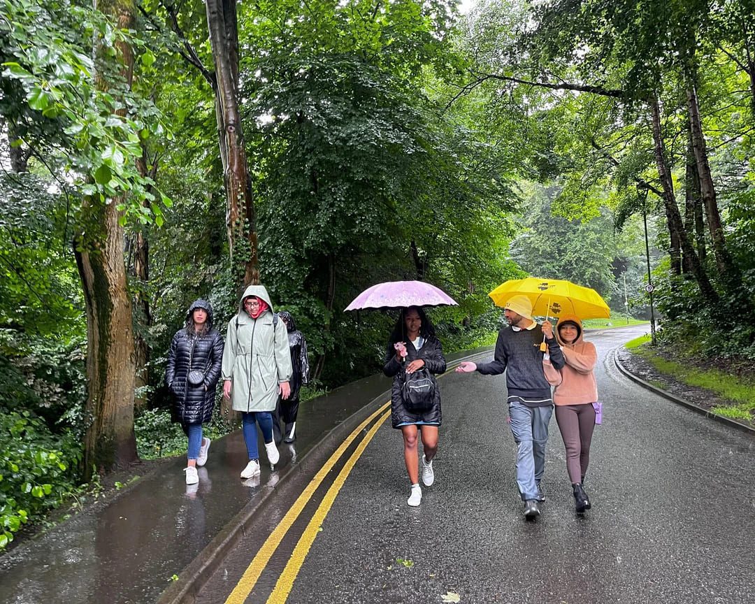 International Student Society members walking with umbrellas on a lush green road in Belfast during a rainy day.